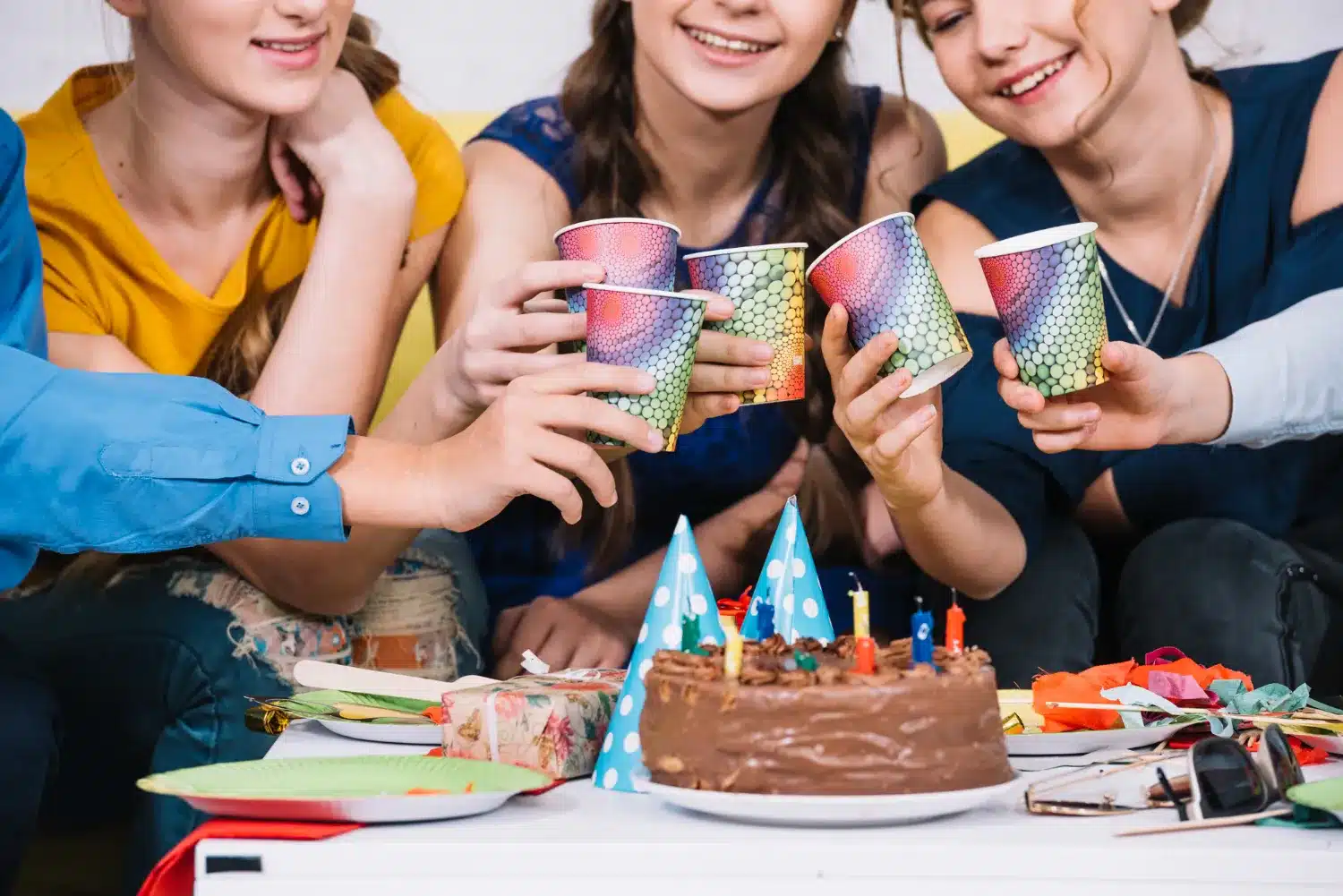 This photo shows three women and the arm of two men celebrating a birthday on a white table. They are crouched around the table. You can't see the girls faces. They're cut off from the nose up so you can only see from the nose down. They are dressed in casual attire. The gentleman have long sleeve blue button down shirts on. You can only see their arms with their hand holding a cup. The women are also holding cups one is wearing a gold shirt to are wearing dark blue shirts. The cups are almost mermaid, looking in styl their paper cups with almost kind of mermaid color designs on them, greenish purple pink. They are sitting over a birthday cake a one tier chocolate, frosted birthday cake with different color, candles, blue, yellow, green and red. There are two blue birthday hats with white polkadots sitting on the table and then just a bunch of random items on the table. There's a pair of sunglasses, a gift wrapped birthday gift a paper plate a few paper plates and what looks like maybe some streamers.