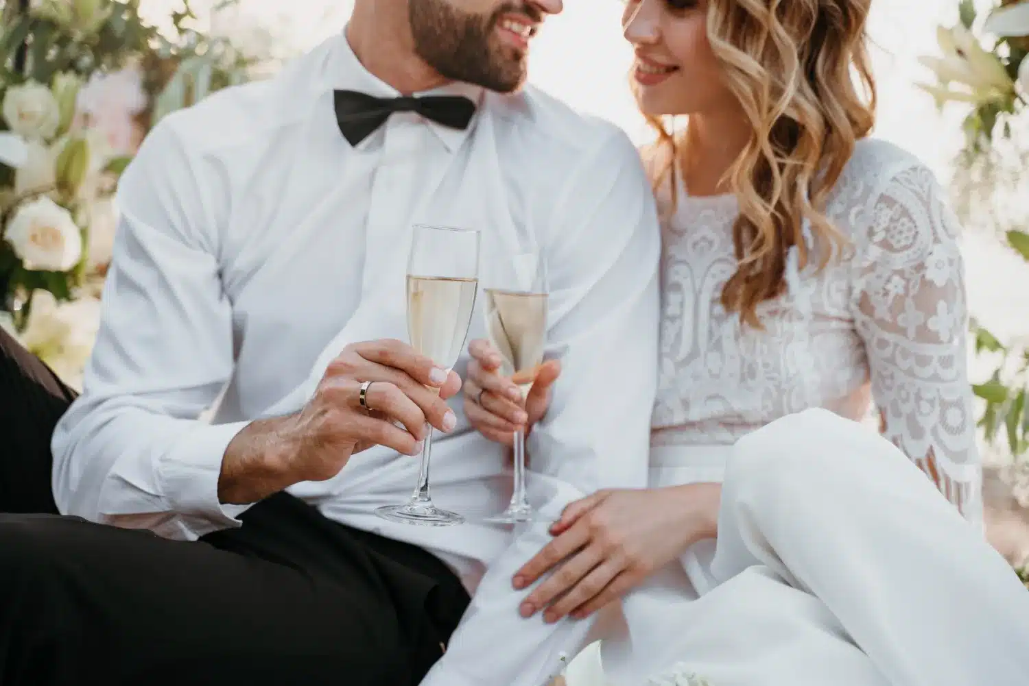 This is a photo of a bride and groom. Their top of their heads are cut off. You can only see from the nose down. She's wearing a white wedding dress. He's wearing a white shirt, black bowtie, and black pants typical wedding attire. They both have a champagne glass in their hand that they are toasting with.