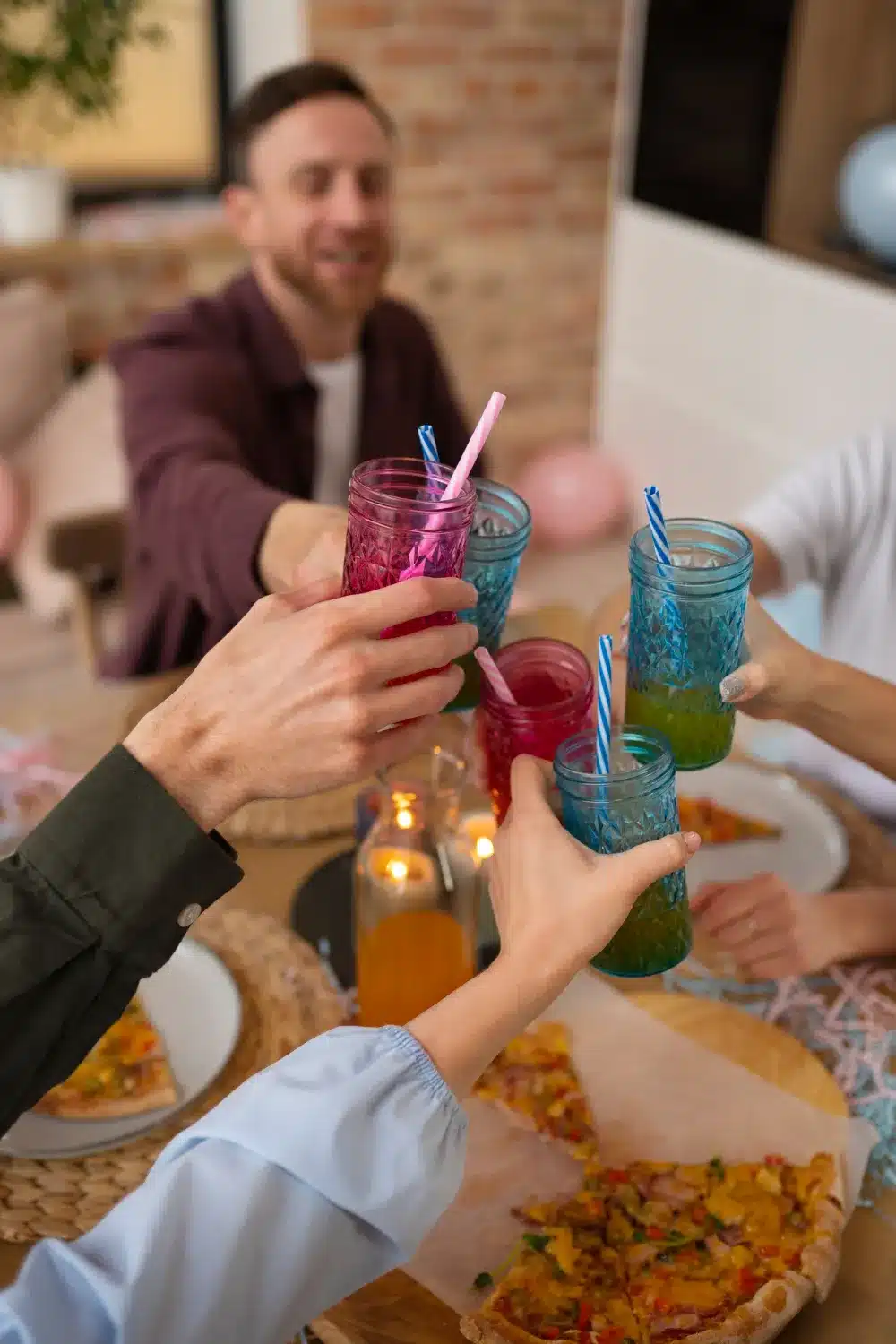 This image shows a group of people cheering with their cup in the middle of a table. The table is light brown wooden color kind of blurry background, kind of blurry table. You can see slices of pizza and a candle. You can see everyone's hands in the middle of the table with their blue and pink glasses with blue and white striped straws and pink and white striped straws, you can see one gentleman in the background his face is blurred out. He has brown hair with a brown beard and a purpleish color jacket over a white T-shirt and then you can only see everybody else's hand and or arm.