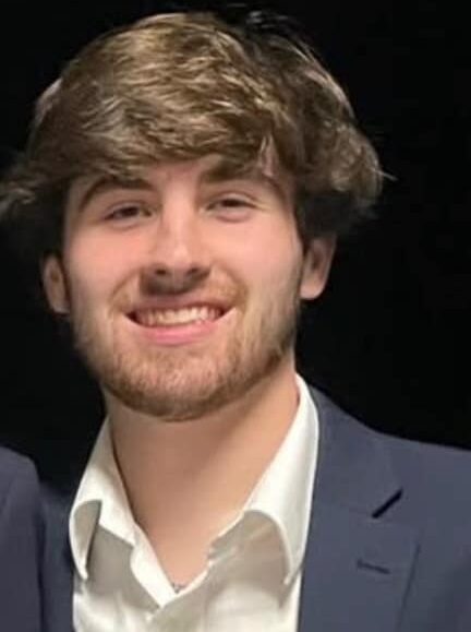 This picture is another team member. His name is Will there is a black background. He's wearing a blue jacket with white colored shirt underneath it. You can only see the very top of the shoulders and neck area. It's a close-up picture of his face. He has lightish brown hai, brown eyes and a beard.