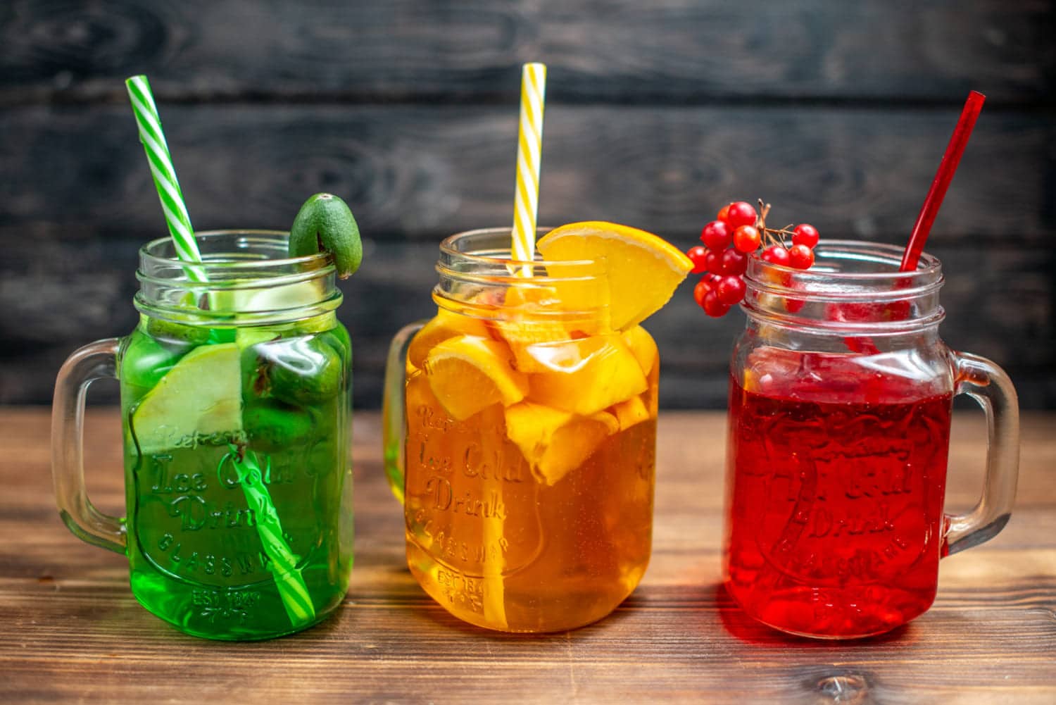 3 class mason jars filled with ice, cut up fruit and topped with sliced fruit and a straw. The drinks are in a row, green then orange, then red. background is a dark grey all and they rest on a brown wood tabletop.