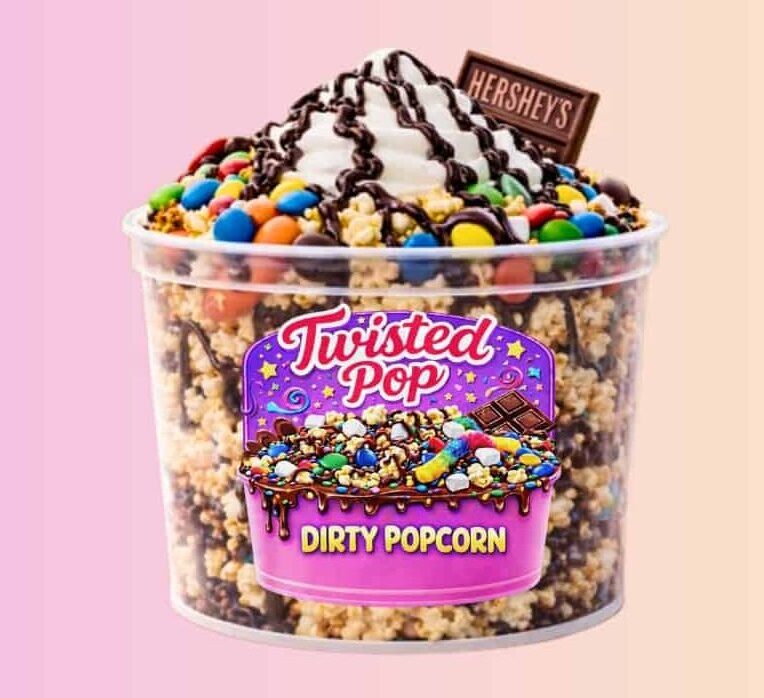 Clear bucket of popcorn with lots of candy mixed in. It has whipped cream on top with chocolate drizzle and a Hershey bar sticking up off the top. There is a super fun purple twisted pop dirty popcorn logo on the bucket with stars and confetti