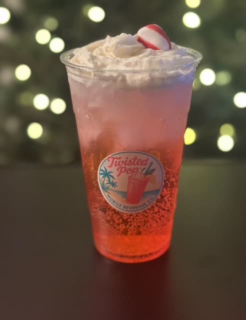 Holiday drink with a blurry Christmas tree with white lights in the background, the drink sits on a black bar top, the drink is in a clear cup with the Twsted Pop logo on it, cup is filled with ice and there is a gradient red drink in it, its more red at the bottom and gets more white on top and it is topped with whipped cream and a red and white mint candy on top