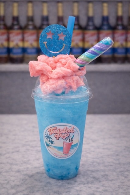Grey countertop with blurred torani syrup bottles in the background, on the countertop there is a clear plastic cup with a twisted pop logo on it, there is a light blue drink in the cup with pink cotton candy on top, and a rainbow colored twist lollipop, with a blue straw and a blue smiley face decor pick on top.