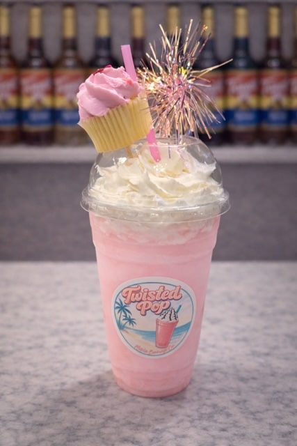 Grey countertop with blurred torn syrup bottles in the background, on the countertop there is a clear plastic cup with a twisted pop logo on it, there is a light pink drink in the cup with white whipped cream topping and a pink frosted mini cupcake with a pink straw and a pink pom decoration.